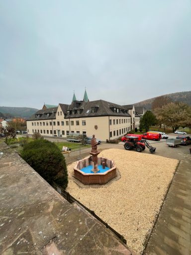 Brunnen mit Statue im Innenhof eines Gebäudes, umgeben von Fahrzeugen und Bäumen.